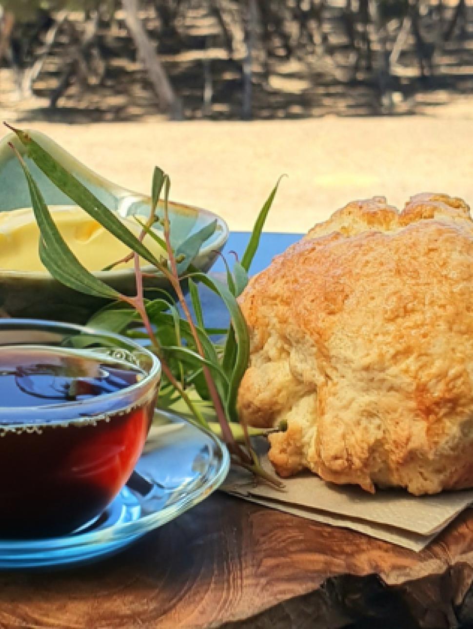 A cup of tea, damper and butter on a wood piece outside