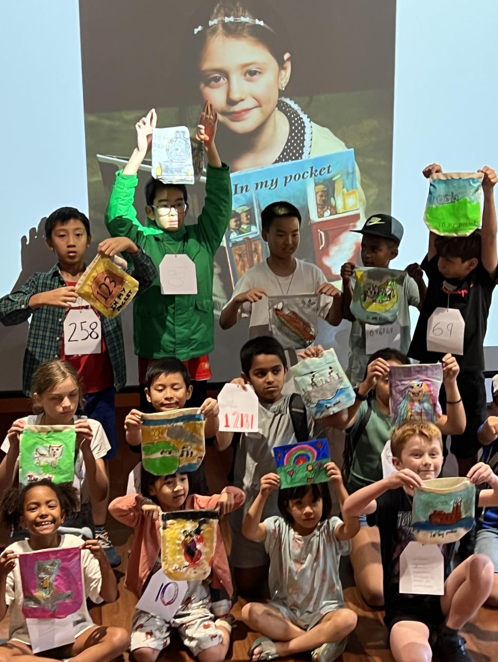 Children holding up artworks in front of a screen