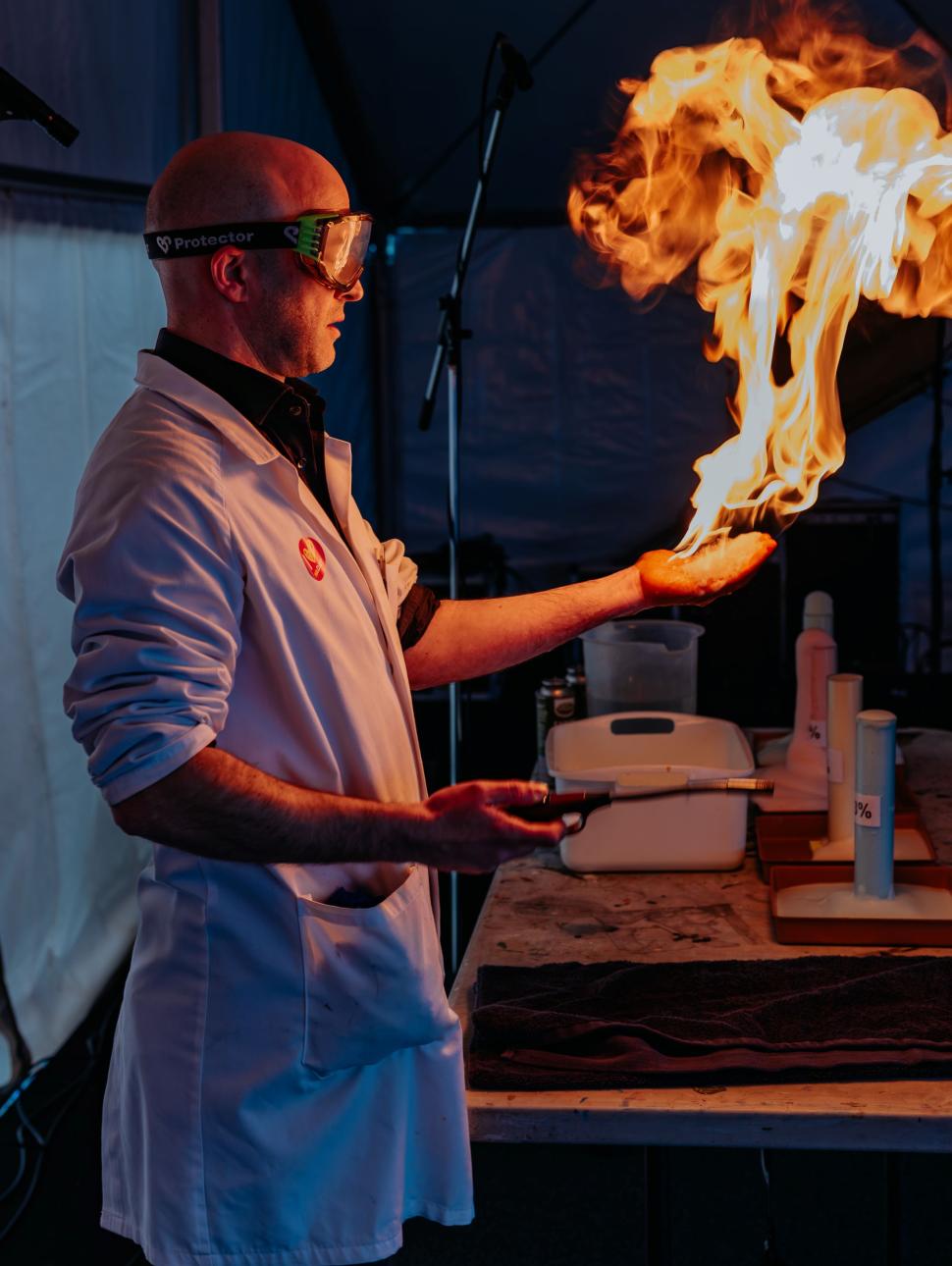 A man wearing safety goggles and a white lab coat with gloves holds a ball of flame, facing to the right