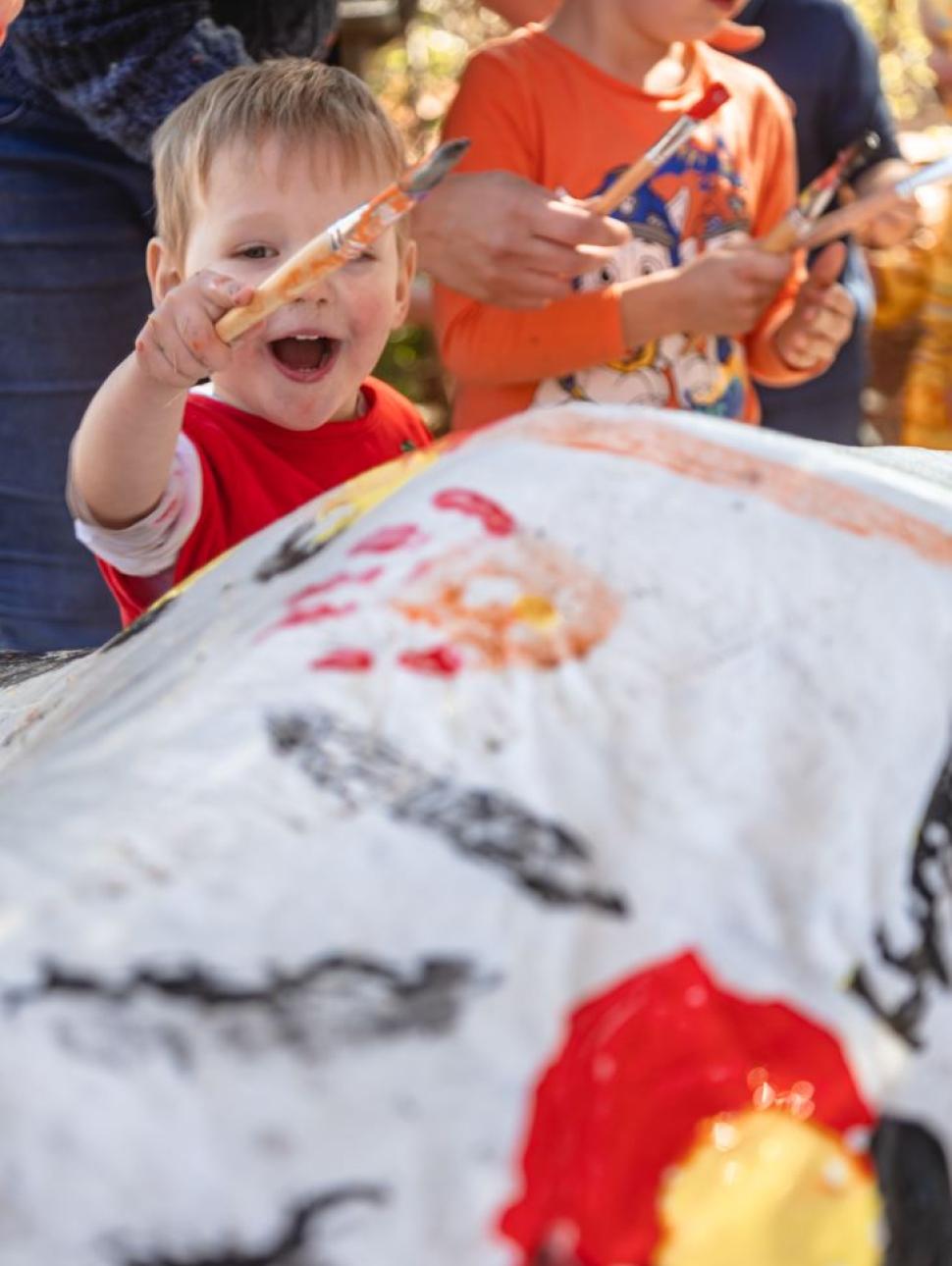 In the photo, a cheerful child is holding a paintbrush with a big smile on their face. They are looking attentively at a sheet adorned with the vibrant colors of the Aboriginal flag.igianl flag