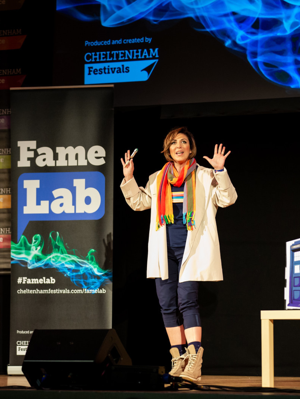 A woman in a white lab coat stands on a stage, exclaiming to an audience 