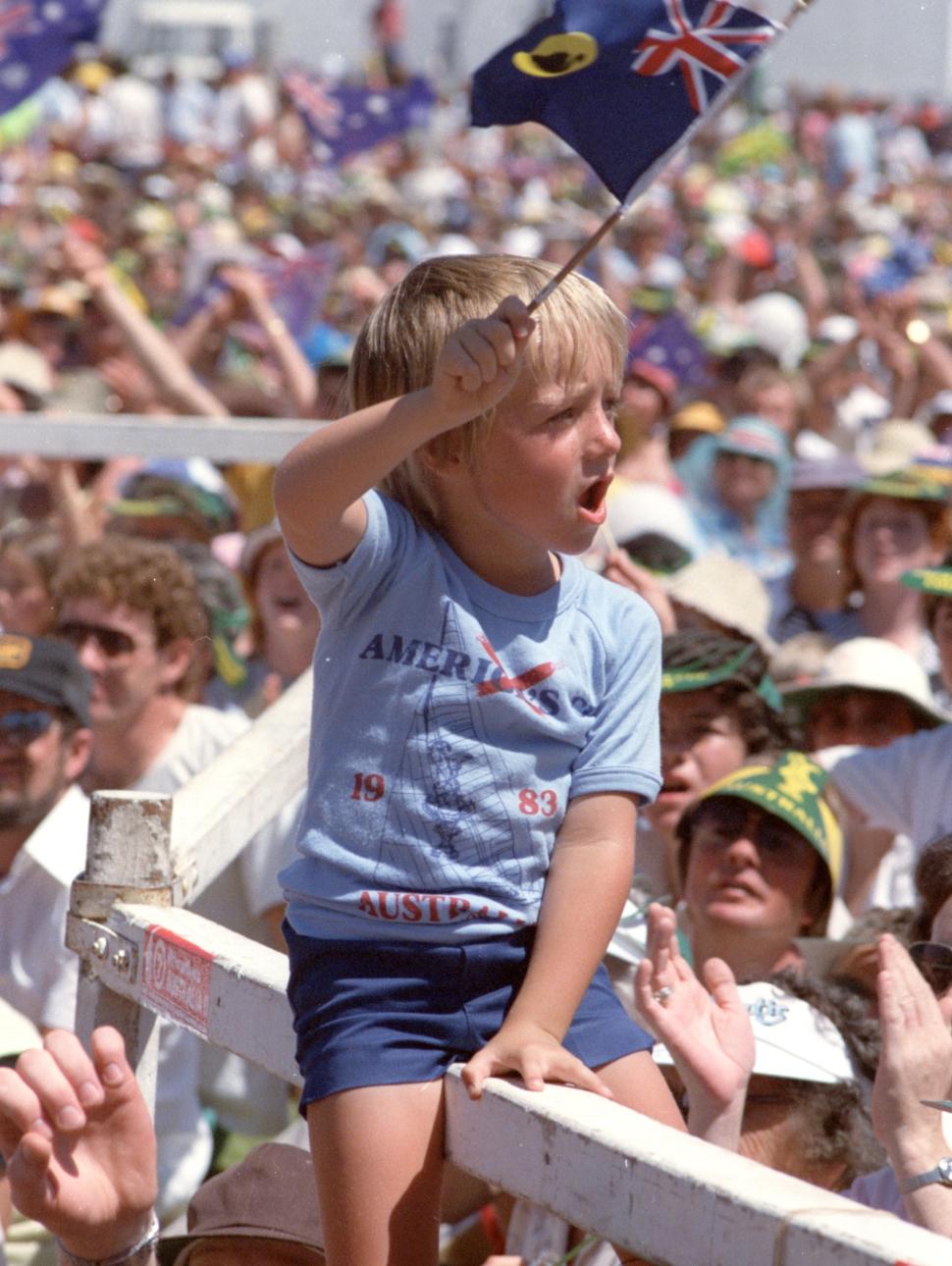 Image of a small boy waving a Western Australian flag in a crowd, attending the Australia II victory concert, Esplanade Reserve, 30 October 1983