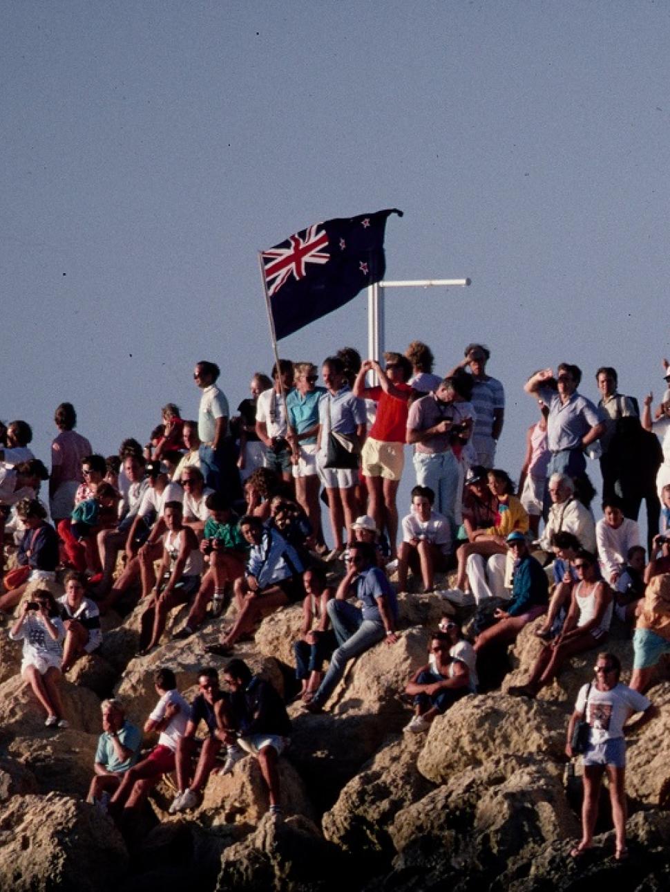 A crowd from 80s era gathers on harbourside rocks with Australian flags in celebration