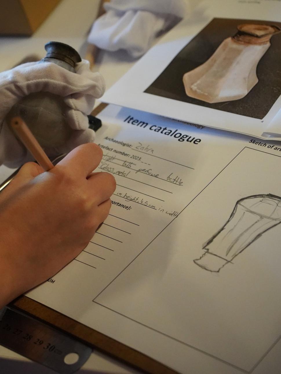 This image shows a close up of someones hand sketching and recording a glass object they uncovered. They are also making some notes on the object to record specific details as an archaeologist would. 