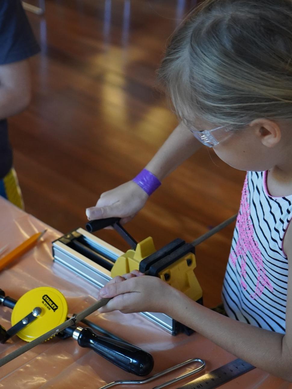 This image shows a young girl using a table vice and hack saw to cut a piece of bamboo. She will be using this to construct a bee hotel. 