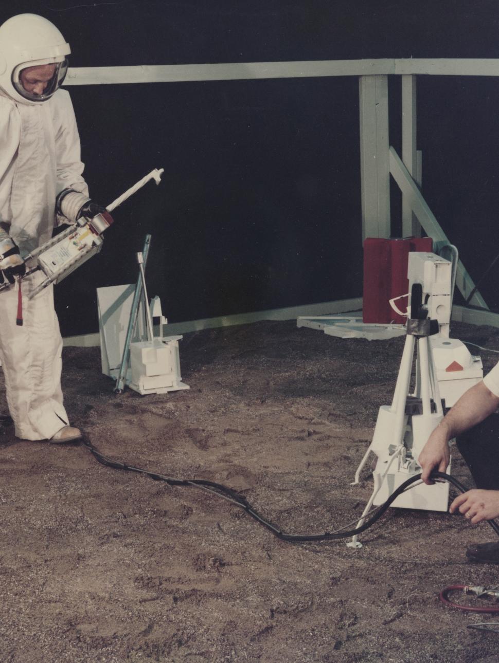 An old image of a man in a large bulky white astronaut suit holding a large instrument with a long spout while another man crouches on the floor, watching