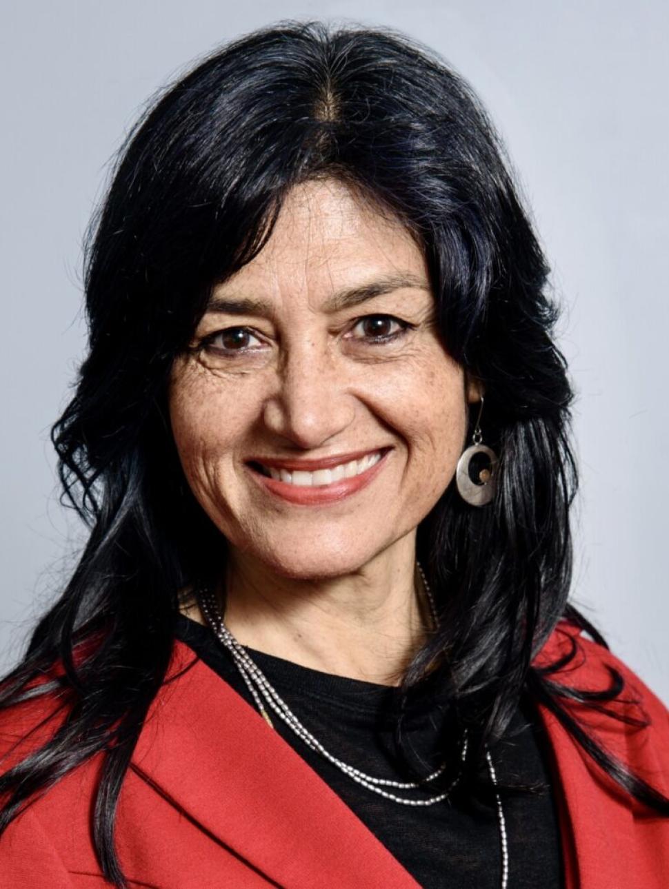 Dr Pilar Kassat is in the middle of the image showing her head and shoulders against a grey background. She is Chilean-Australian, she has long dark hair and brown eyes. She is wearing a black top under a bright red blazer and has large silver earrings and a beaded necklace. She is smiling widely.  