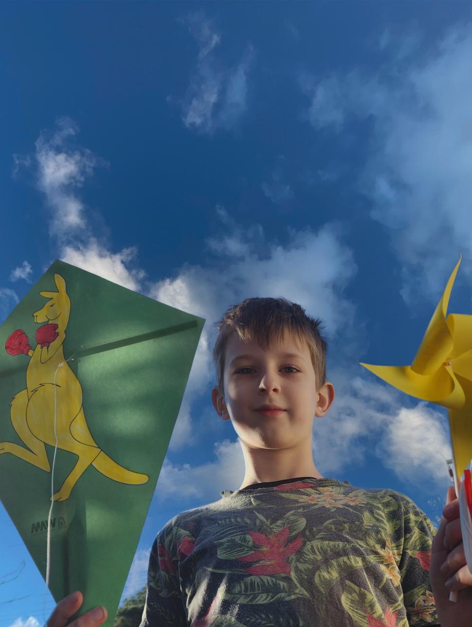 A child of approx 10 years is smiling and holding green kite with a yellow kangaroo and in the other hand they hold yellow and green spinning wheels. A blue sky is in the background