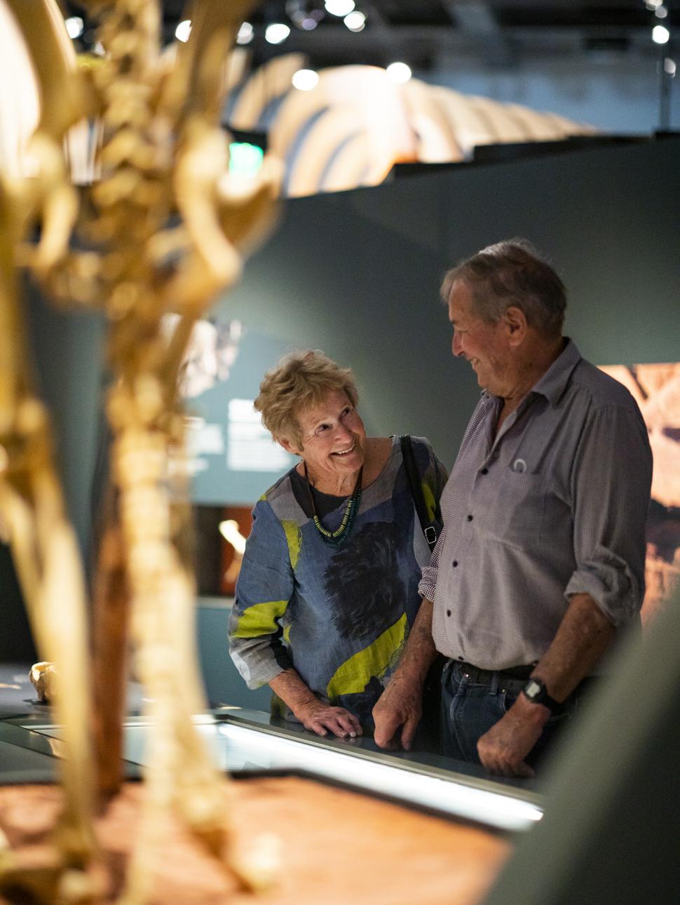 Two senior individuals studying a skeleton fossil together, with the fossil in the foreground and a gallery setting in the background