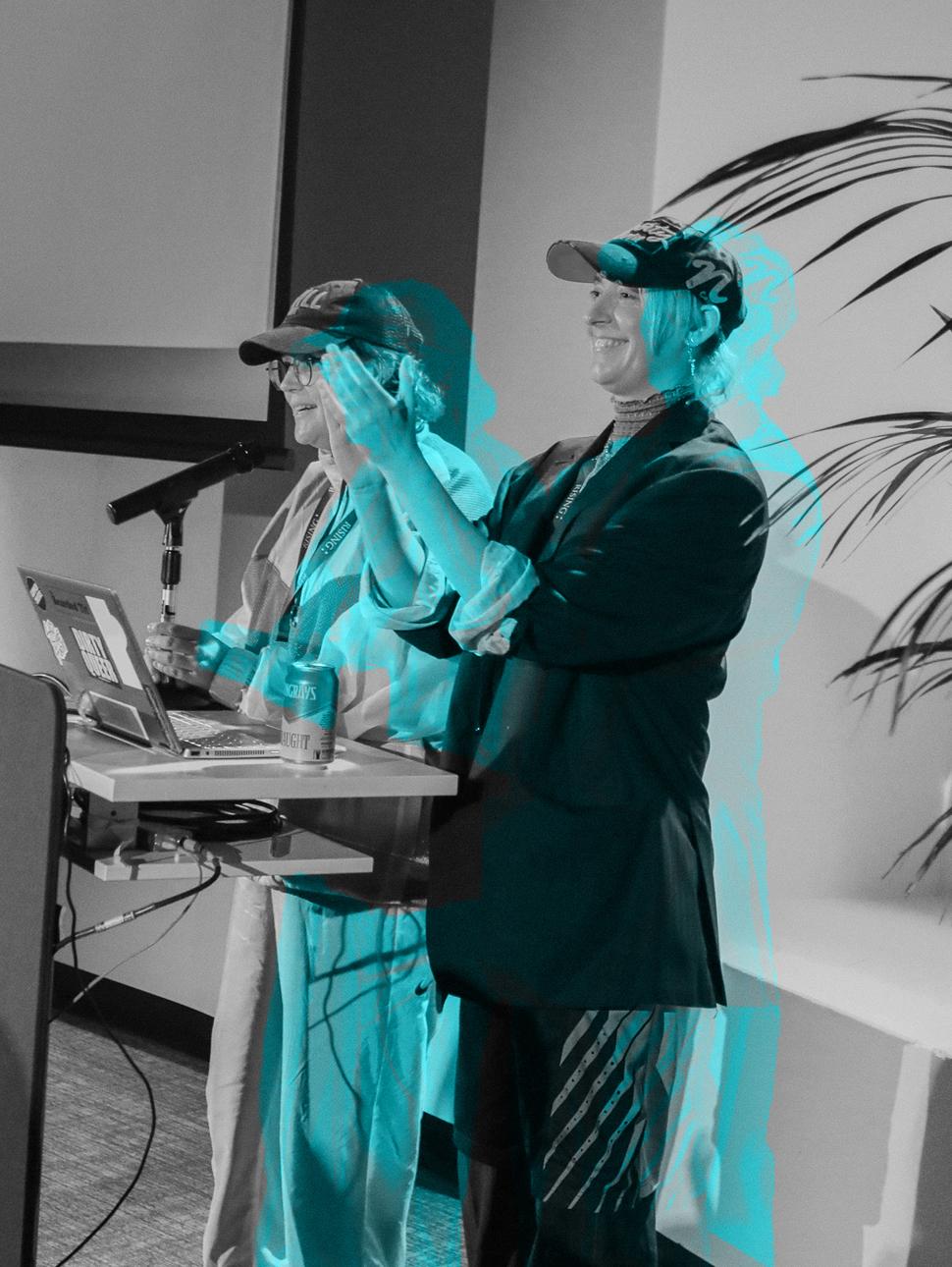 In a theater setting, two hosts are standing on a stage. In front of them, there is a laptop displaying a black and white image, which is overlaid with a vibrant and bright blue visual treatment.