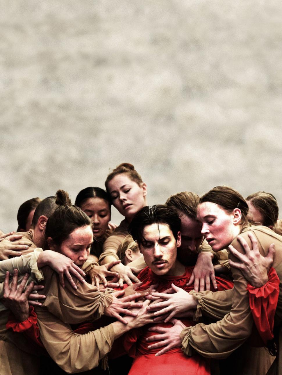 A group of dancers in brown textural floaty costumes gather around the main dancer who is dressed in a bright red costumefo the same design. The dancers hand are all interwoven as they clutch at each other creating a mass of bodies.