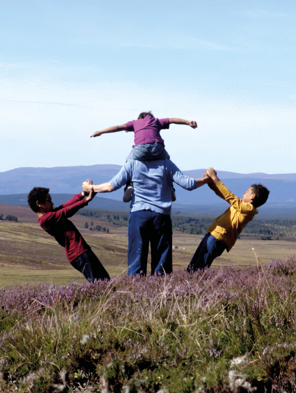 A mother and three children stand and look out onto the moor. One is on her shoulders, the other two hold an arm each and lean away from her.