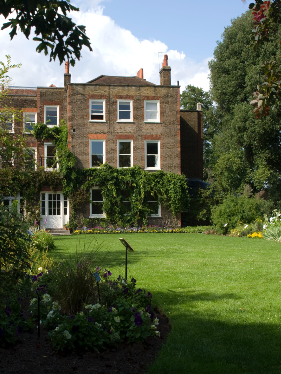 Flat green lawn area surrounded by English garden and tall trees leading towards a three story old brown brick large house