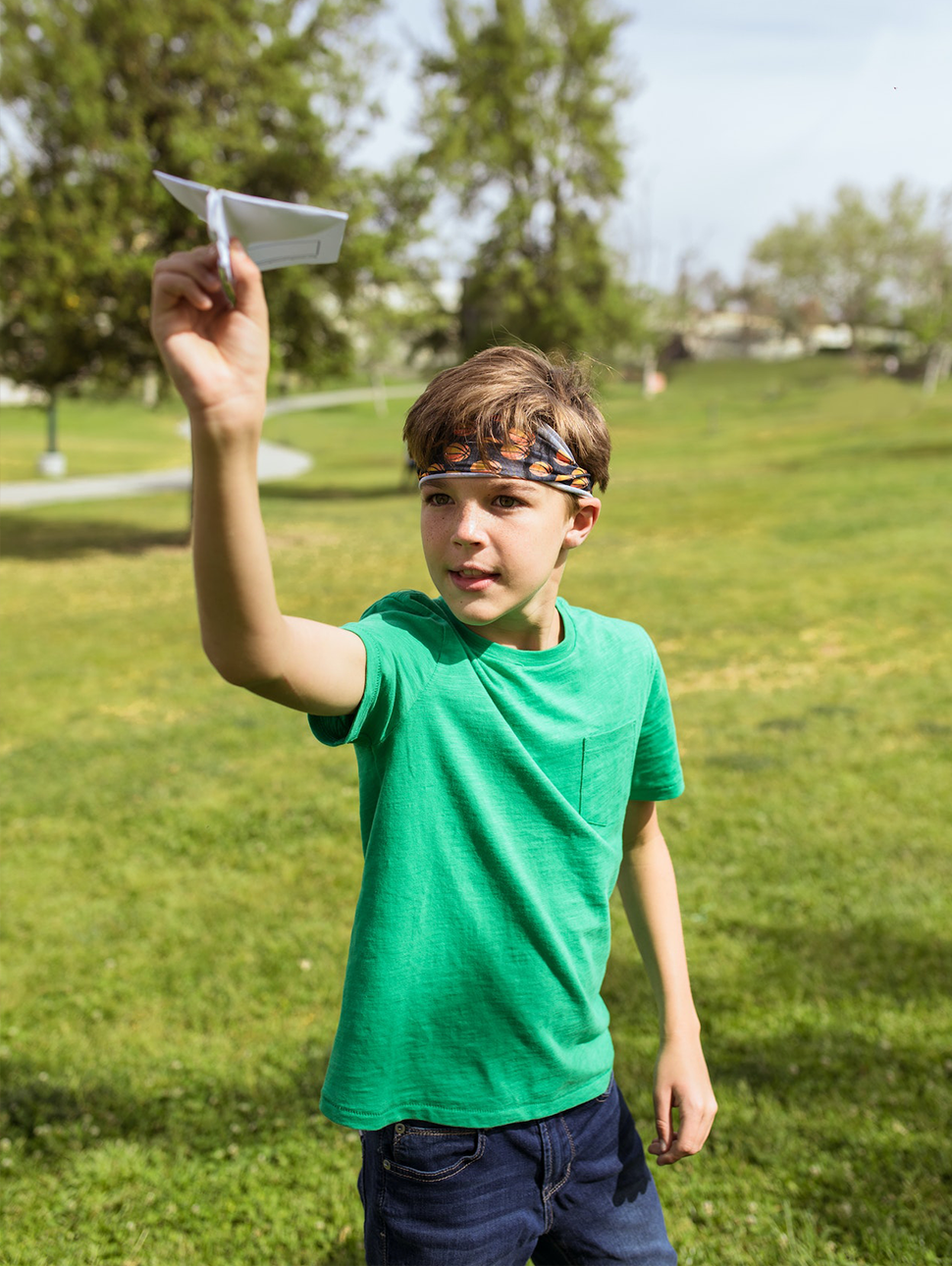 a child stands in a park with a green tshirt on and a headband. they hold a paper plane in one hand and aim to throw it