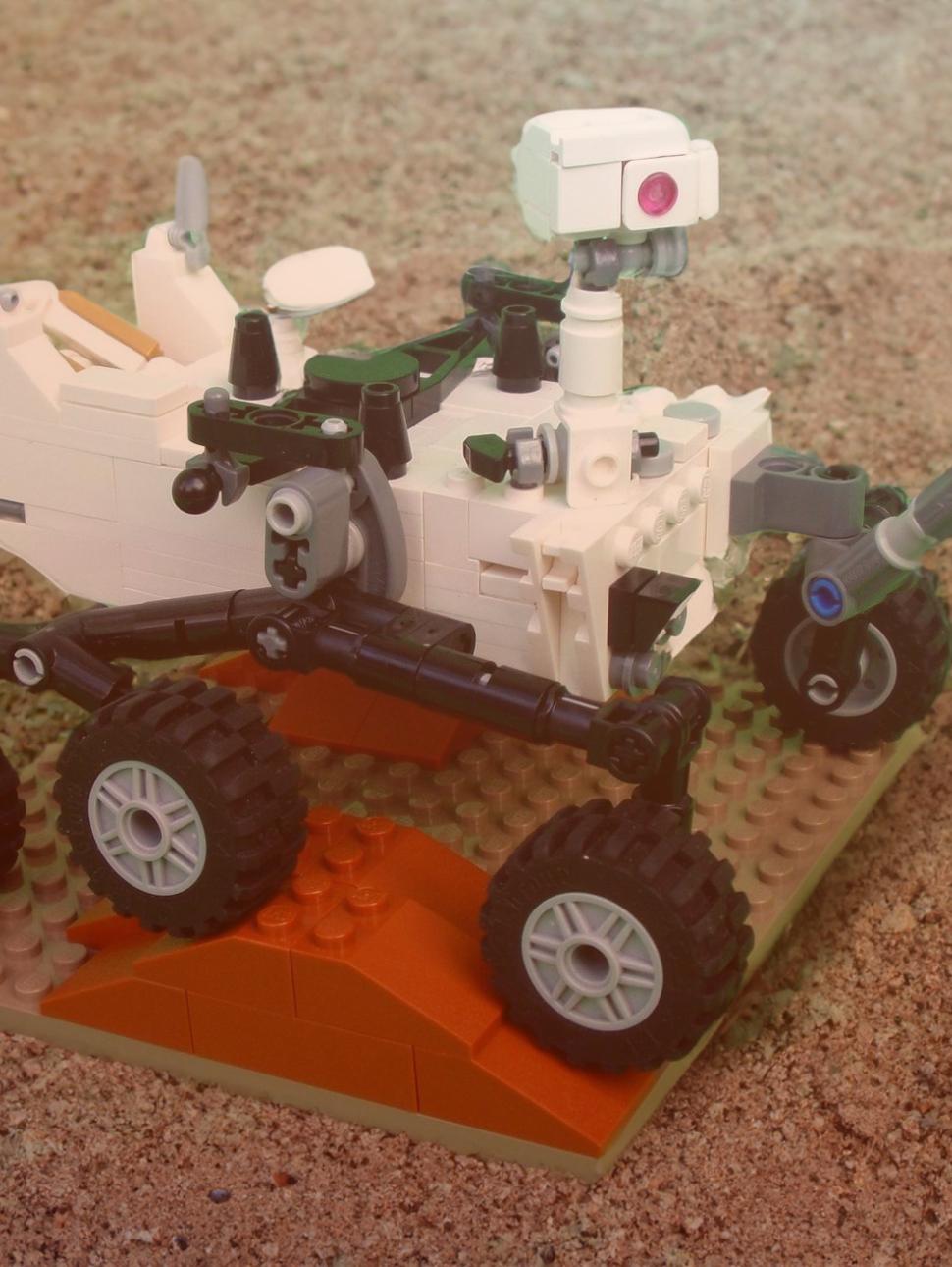 a small planeta lander is made out of lego block pieces mostly white pieces and has six sets of wheels driving over terrain which hs rocky and sandy. there is a slightly green and red tinge to the picture