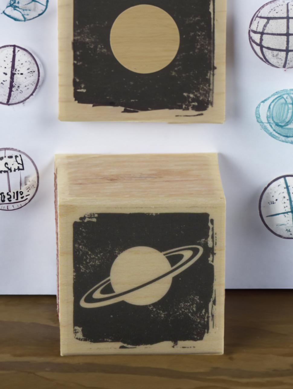 a woodern block stamp of saturn with another planet orb stamp just behind it with a page of a few planet stamps