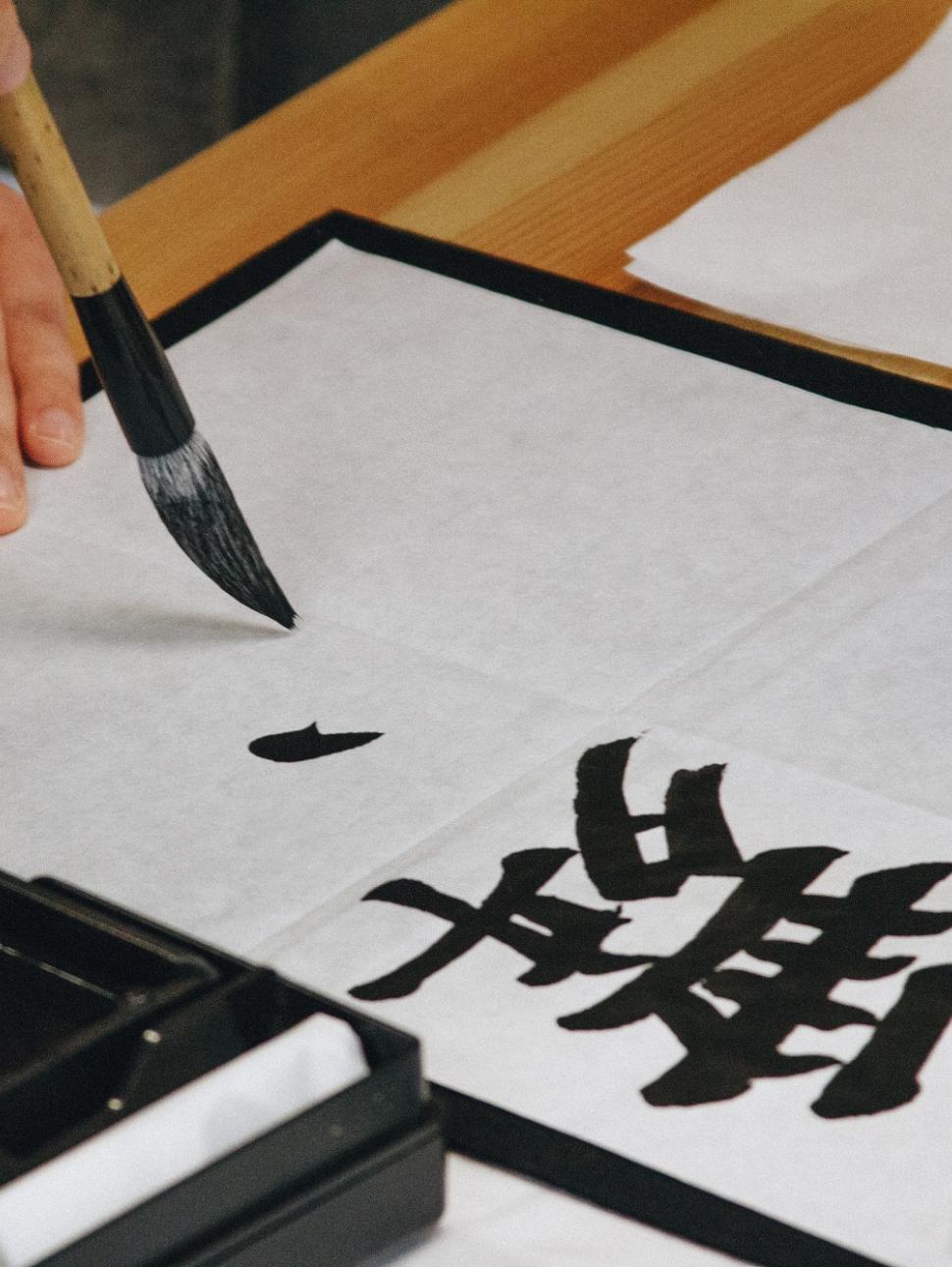 A hand holds a paintbrush which paints with black ink on a white page. the writing is calligraphy of Japanese language characters