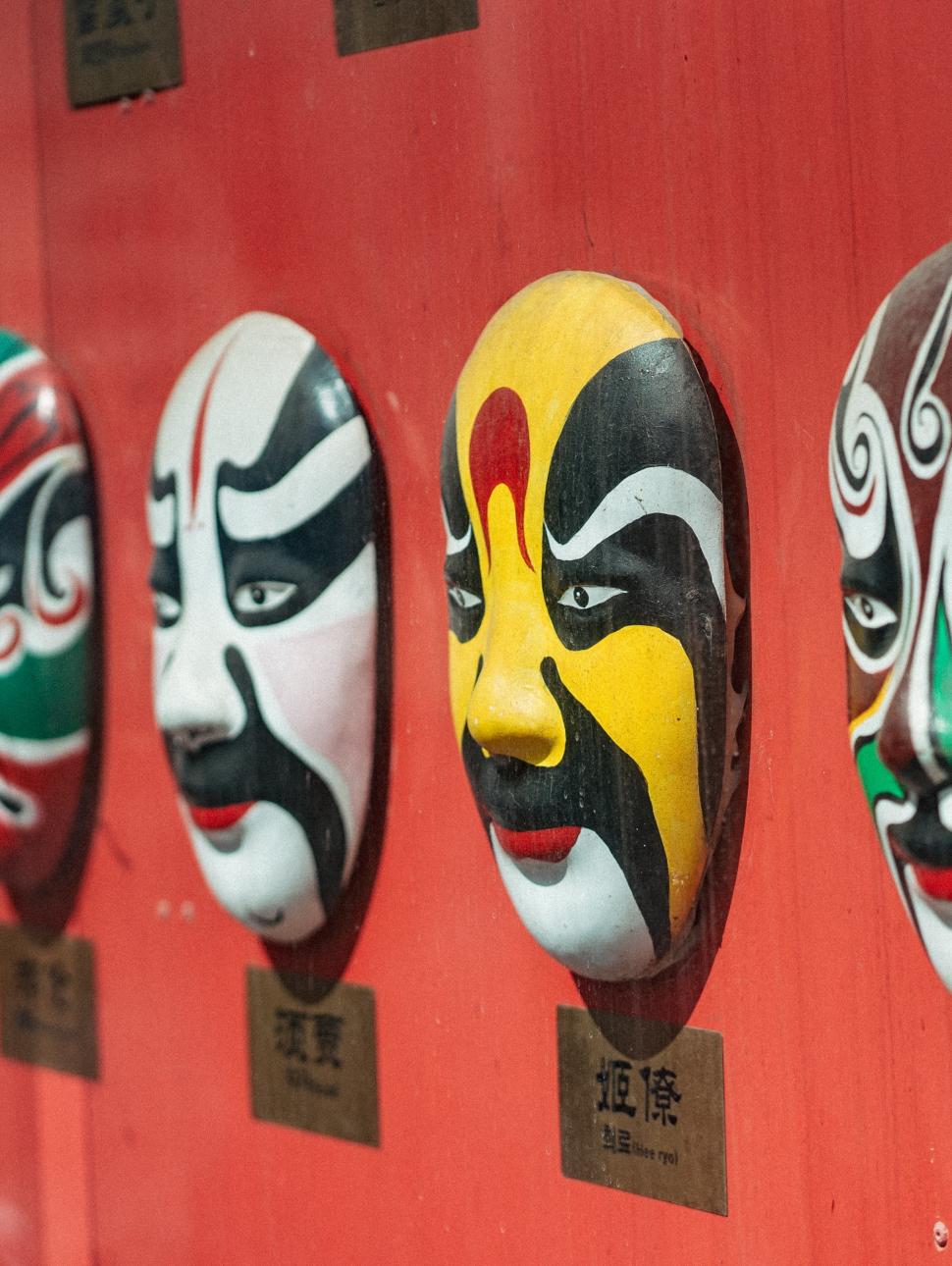 a row of highly deecorated full face masks are on a red wall displayed. each mask has a white colour base and decorated uniquely with many colours