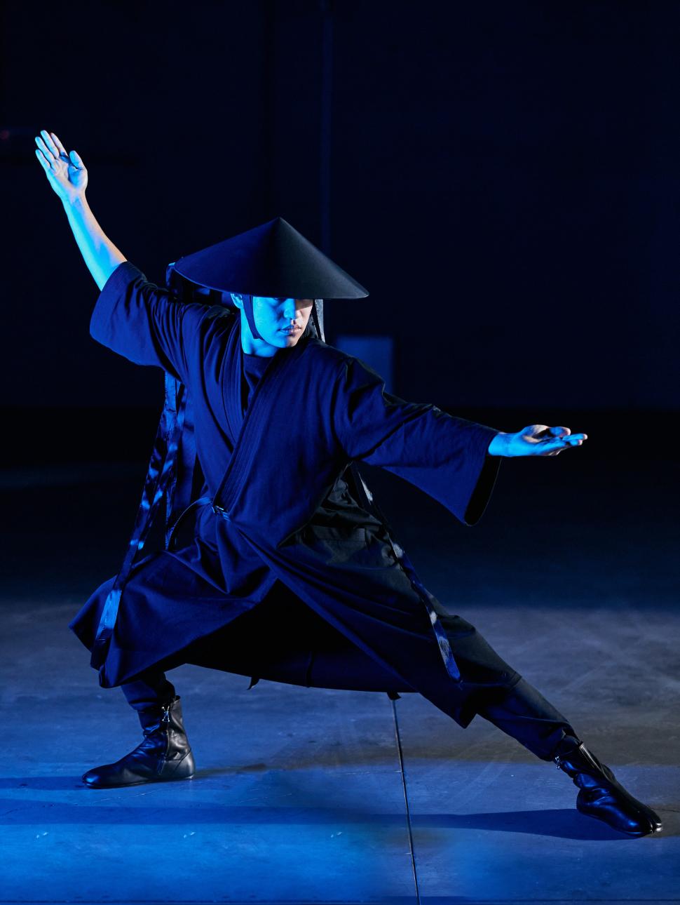 an adult person dressed in all black flowing clothing and hat poses with arms outstretched in opposing directions in a dark room