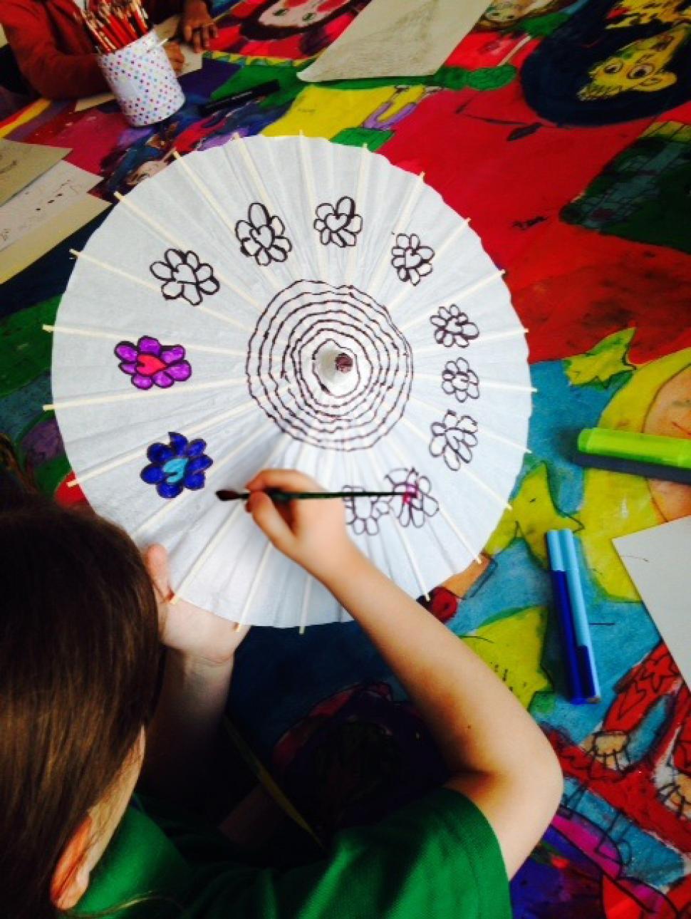 an overhead shot of a young child 10 years old painting a white paper parasol umbrella with bright paints 