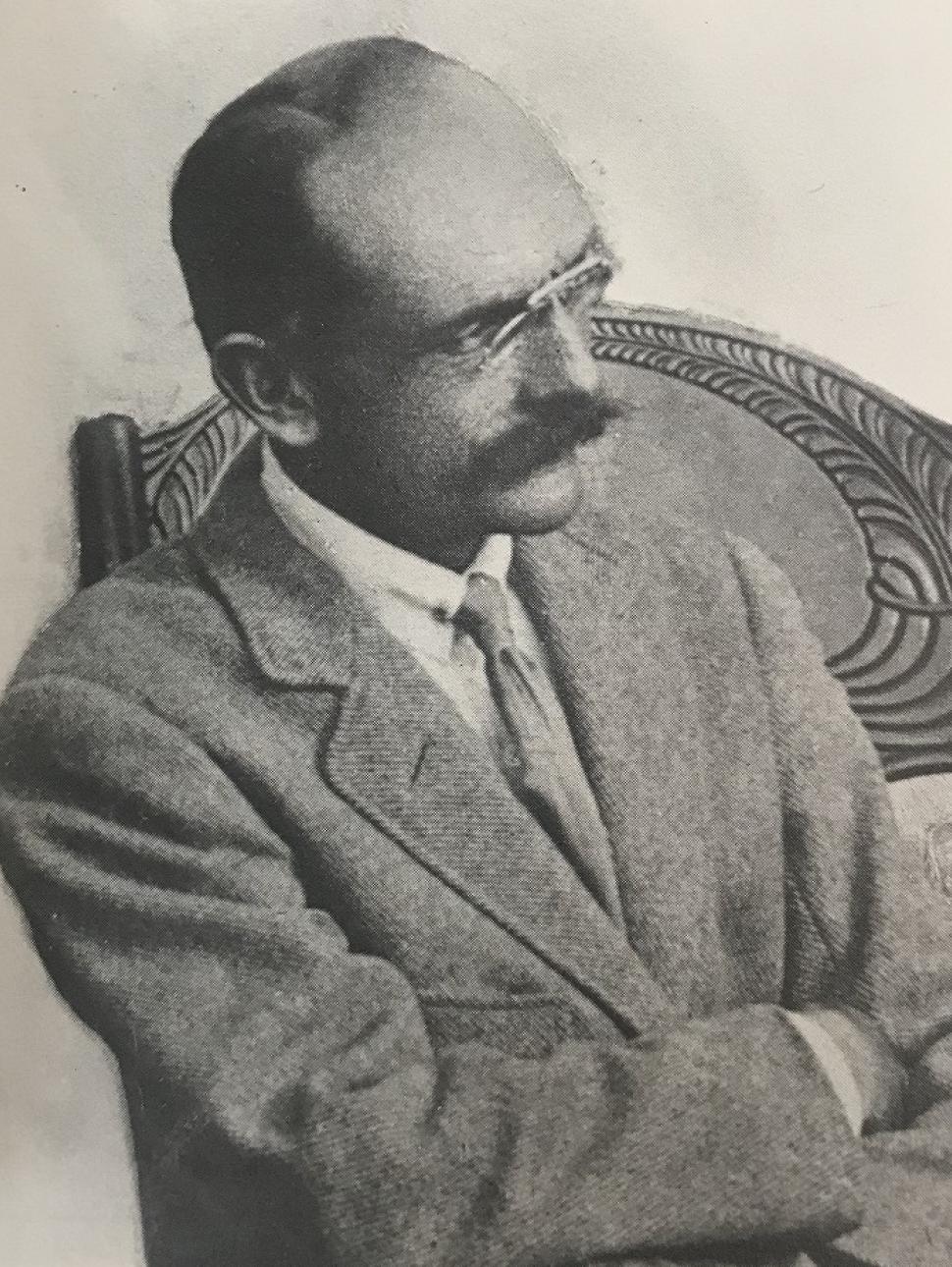 Black and white photo of balding man with a bushy mustache seated on a wooden chair wearing a suit and tie with his arms folded.  Wearing glasses he is looking away from the camera.
