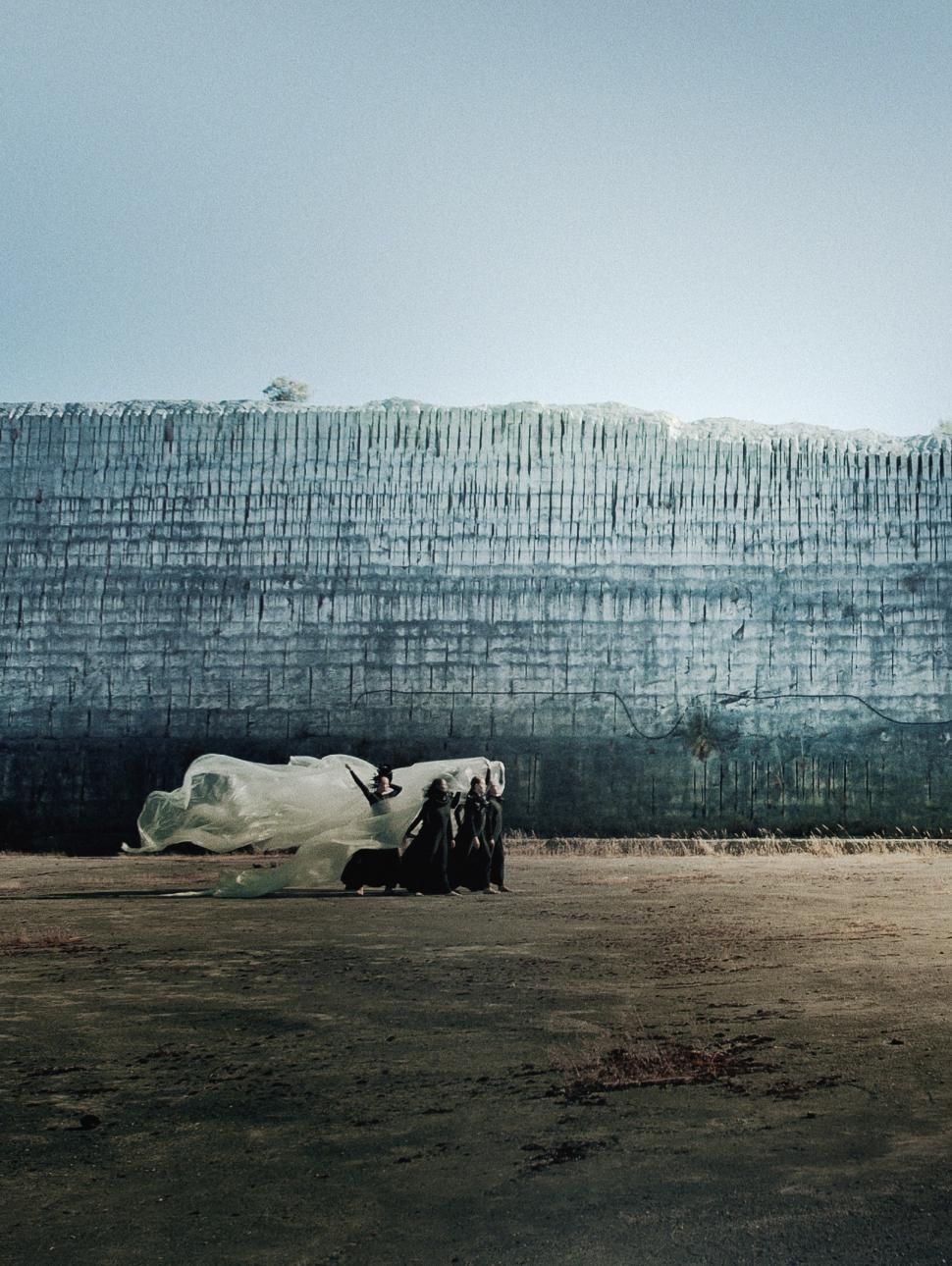 Four dancers wearing long black dresses walk in a procession across a barren space in front of a high blue concrete wall. The dancers hold aloft a large white piece of fabric billowing out behind them in the breeze. 