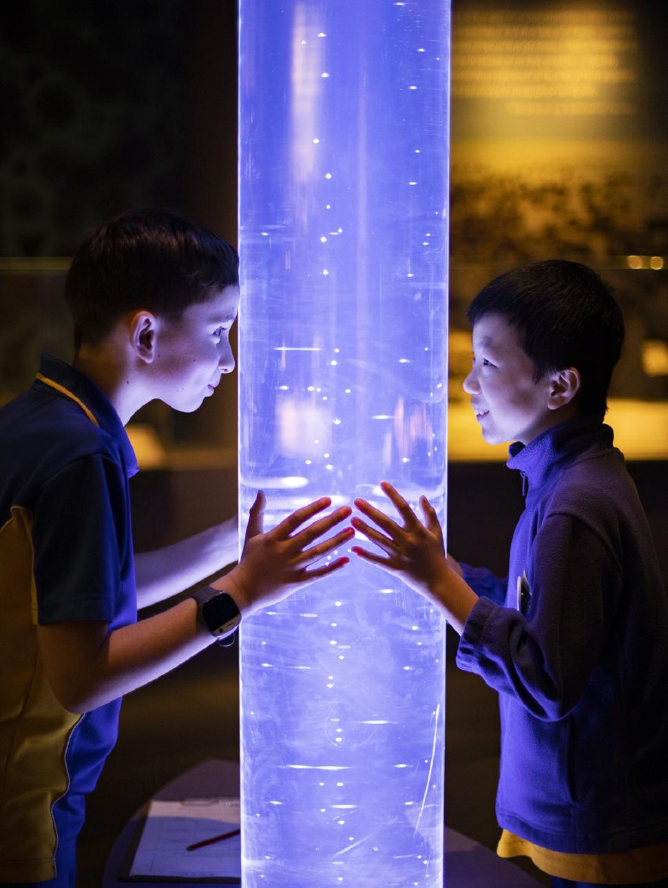 The image shows tow boys standing either side of a large plastic tube filled with a liquid with air bubbles going through it. It looks purple in colour