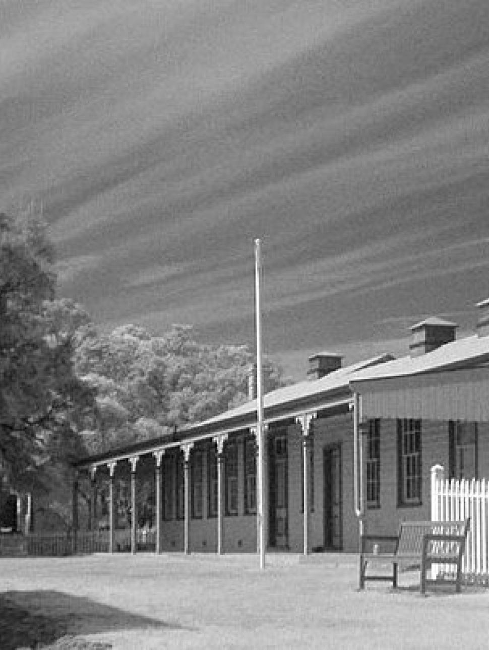 Black and white photos of an old style weather board clad building with a front verandah, and next door is a white picket fence.  On the other side of the building are tall trees.