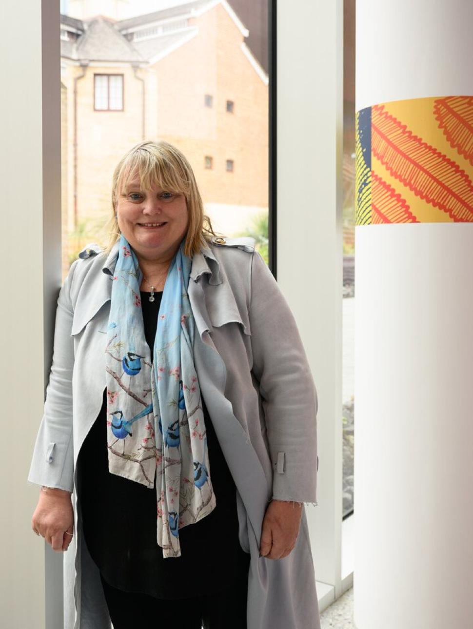 Headshot of Kliti Grice standing next to a pillow with Indigenous art 