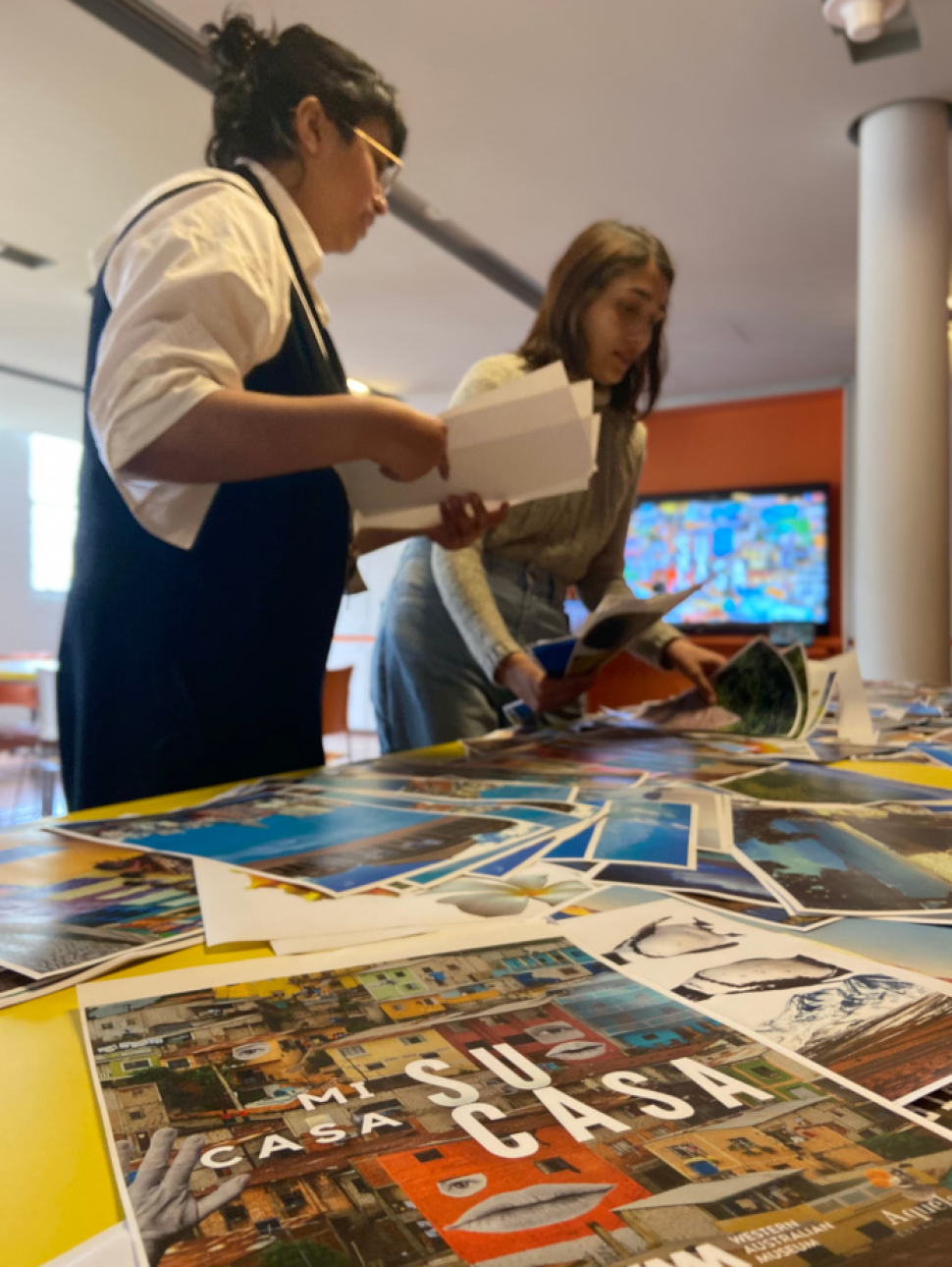 Two women of Latin American heritage stand at a table in conversation. Art work and collage materials are spread across the table. In the direct foreground is a poster for the exhibition 'Mi Casa Su Casa'