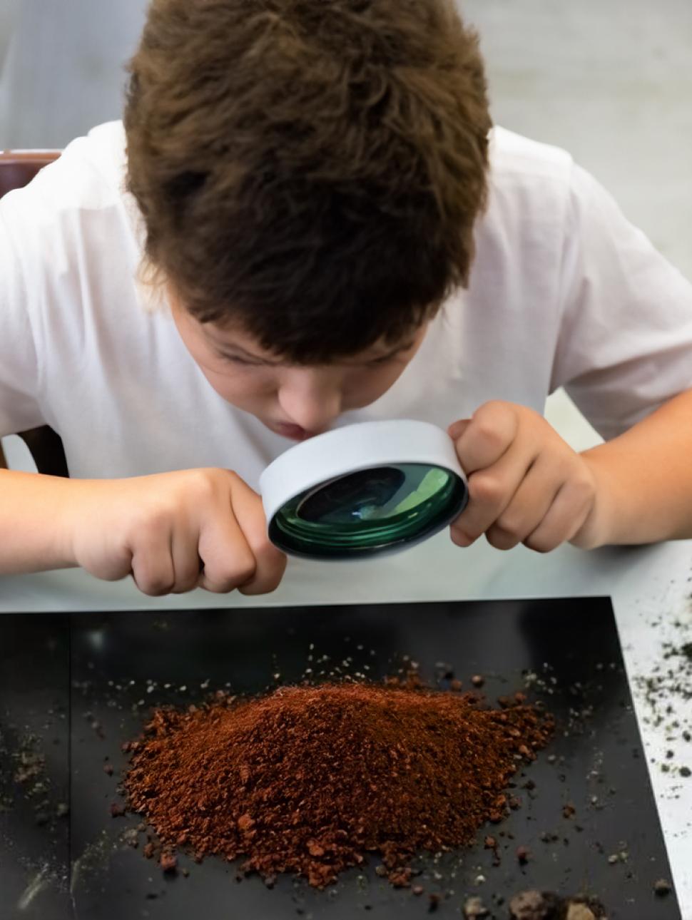 a 12 year old child looks at some soil on black surface through a magnifying glass