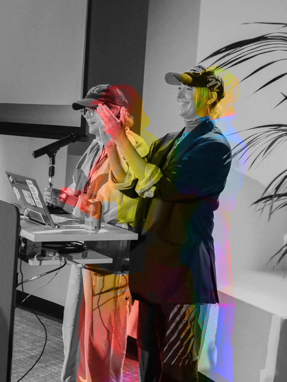 Two presenters enthusiastically engaging with the audience while gathered around a desk with a laptop, set against a black and white backdrop with a vivid, colorful filter applied.