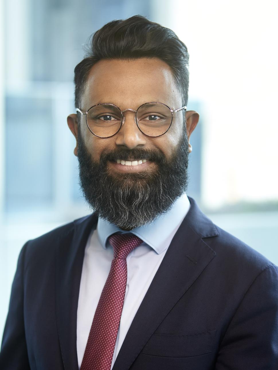 headshot photo of Mr James Jegasothy. James is in the forefront of the photo, he has black hair, beard and moustache, brown eyes and a friendly smile. James wears glasses and is wearing a blue suit with s red tie.