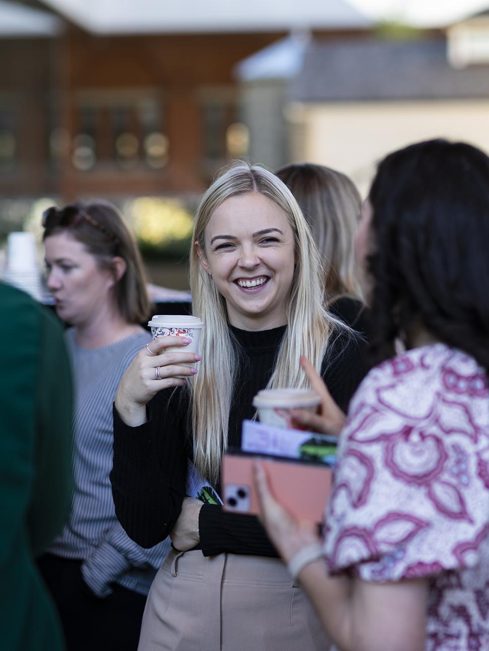 A young blonde woman stands smiling holding a coffee cup