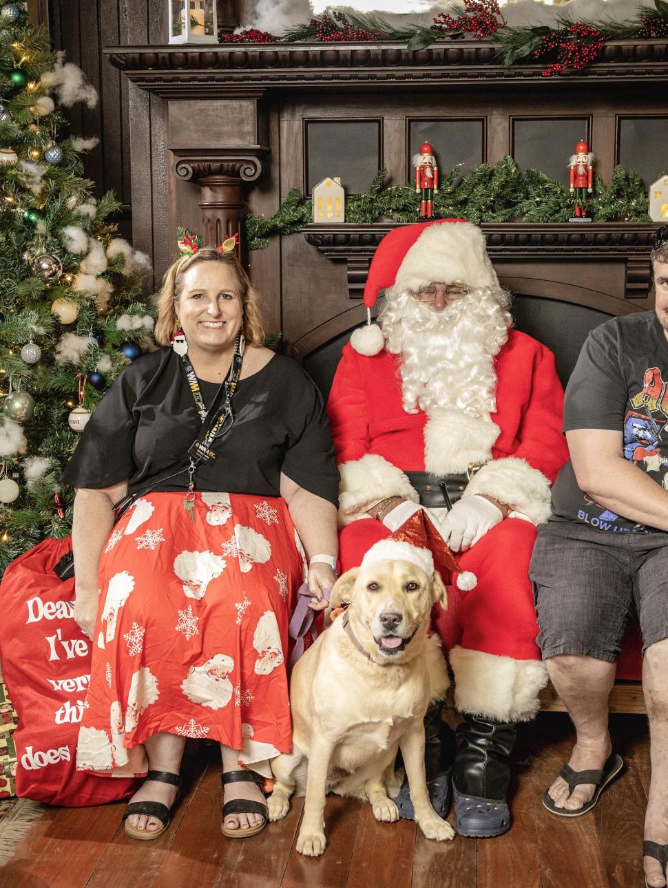 two people sit in a large lounge chair next to a person dressed in red as Santa. Two pet dogs sit alongside this group of people among a Christmas festive setting