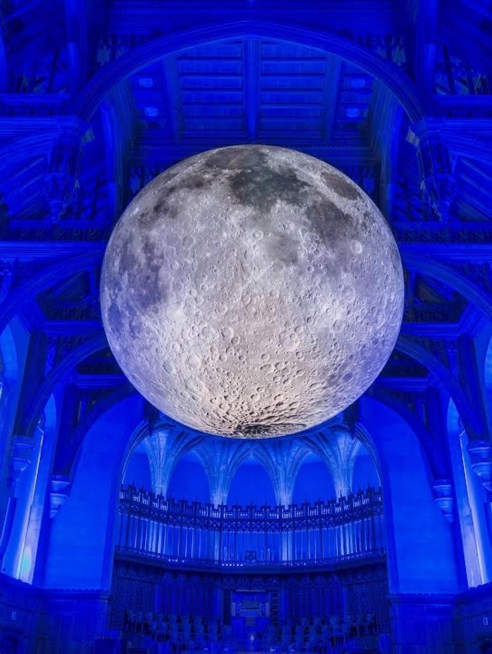 A large moon hangs in the middle of the Great Hall at the University of Bristol UK, where it is lit in a dazzling white against the atmospheric blue lighting that fills the historic building 