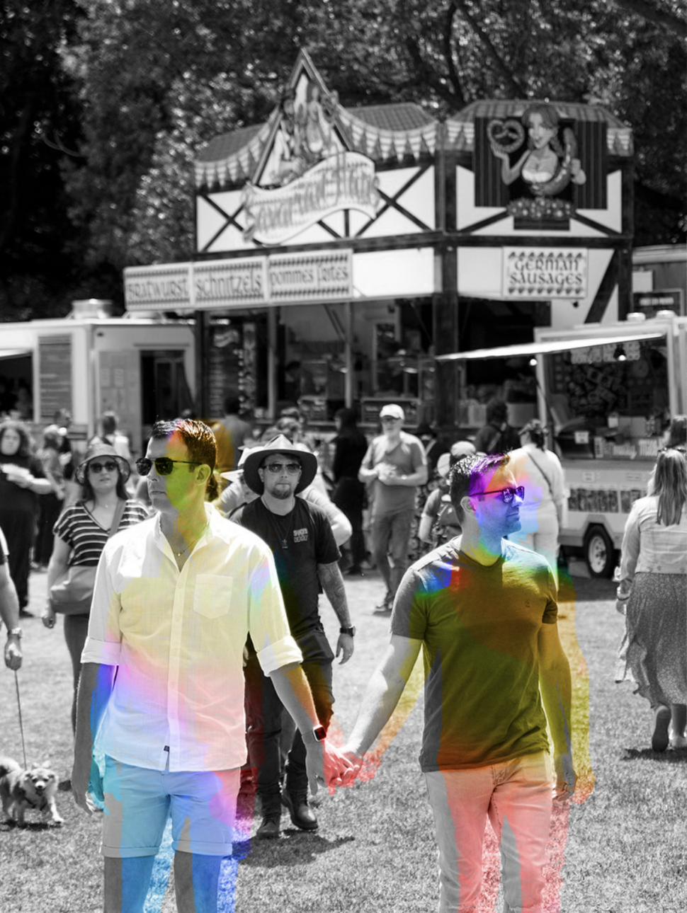 A black and white image of two men wearing casual clothing and sunglasses holding hands at a kind of community fair event. The men are highlighted with a technicolour effect that sets them apart from the black and white background