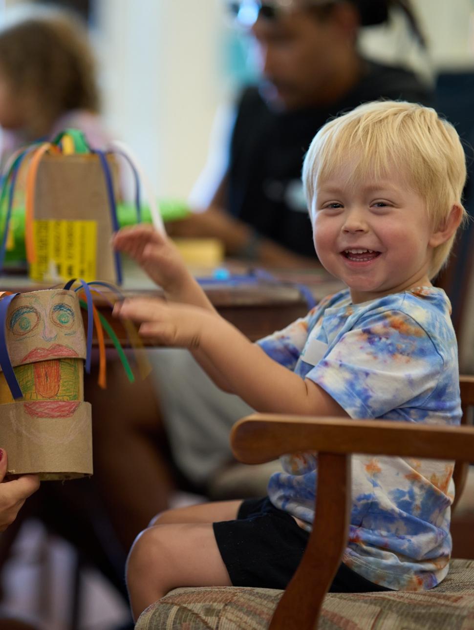 A young blonde child in blue tie dye t-shirt smiles at camera whilst holding puppet made of cardboard, with an adult woman knelt next to him, looking happy