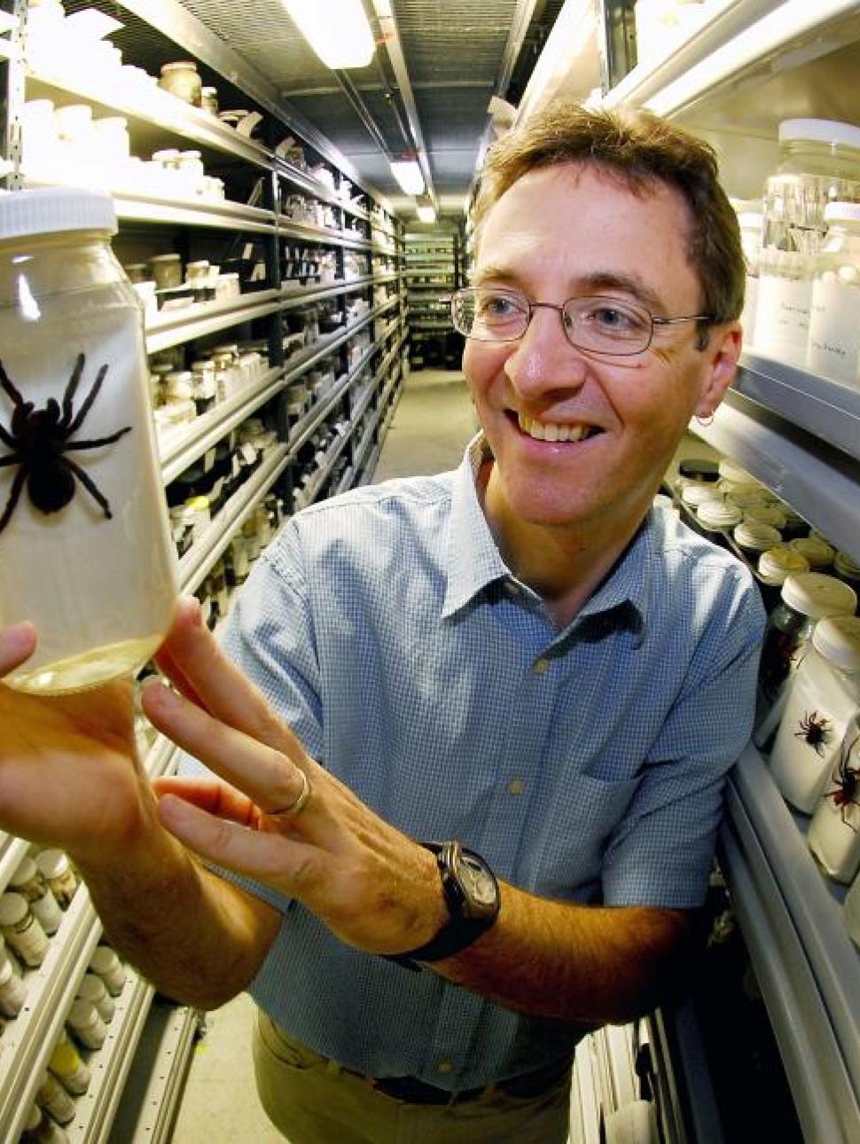 Dr Mark Harvey wears a blue button up shirt, tan trousers and thin framed glasses, and smiles as he holds up a specimen jar containing a large black spider. He stands in a collections facility surrounded by jars of spider specimens