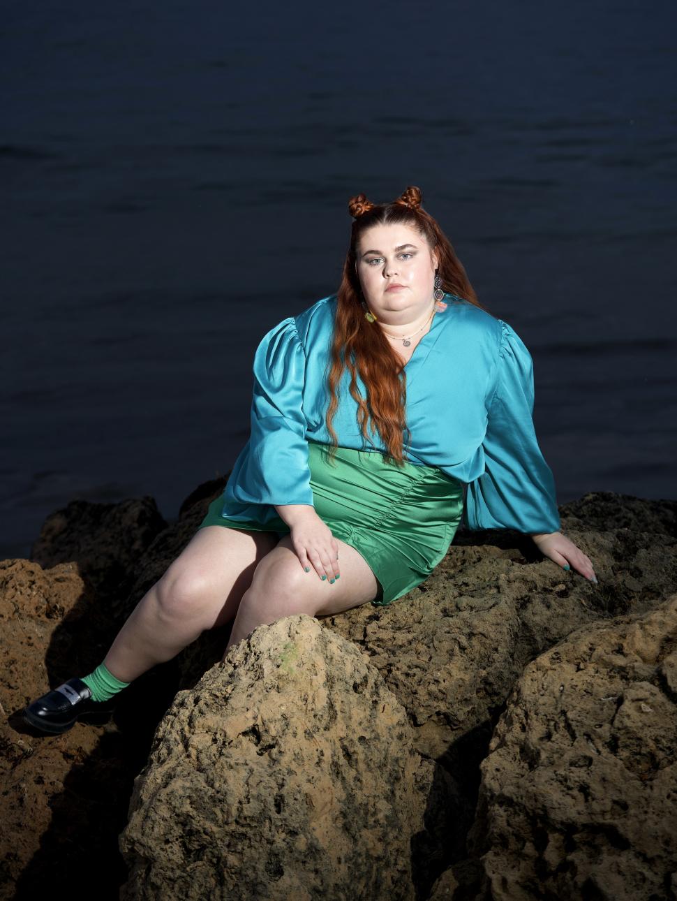 A red haired person with an aqua blue top and emerald green pencil skirt sits on a rock looking seriously into the camera. Brightly lit against the dark nighttime ocean behind her, her outfit appears iridescent in the light