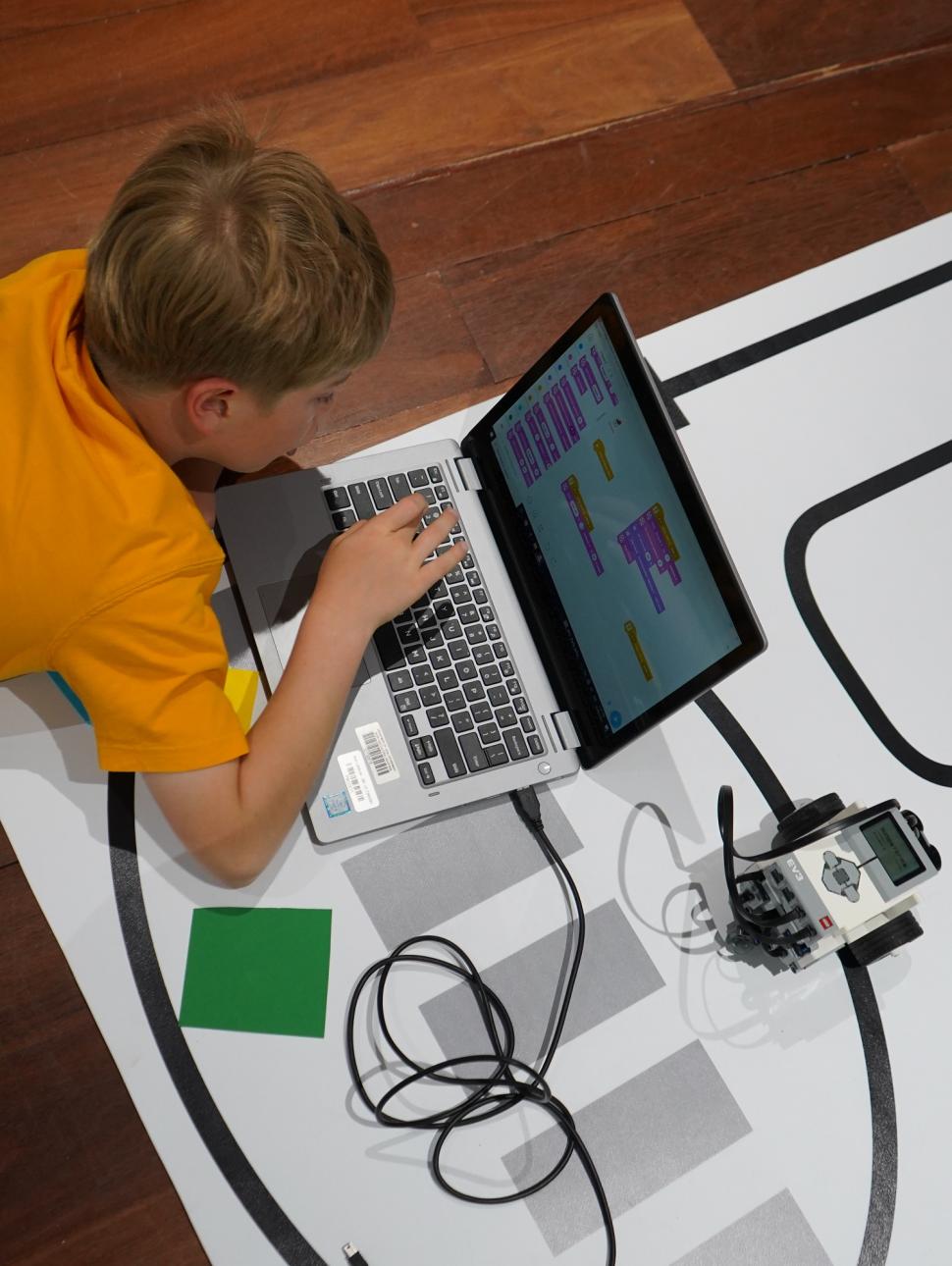 This image shows a burds eye view of a child  on the floor using a laptop computer  to program a  robot to follow a pathway printed on black and white a floormat. 