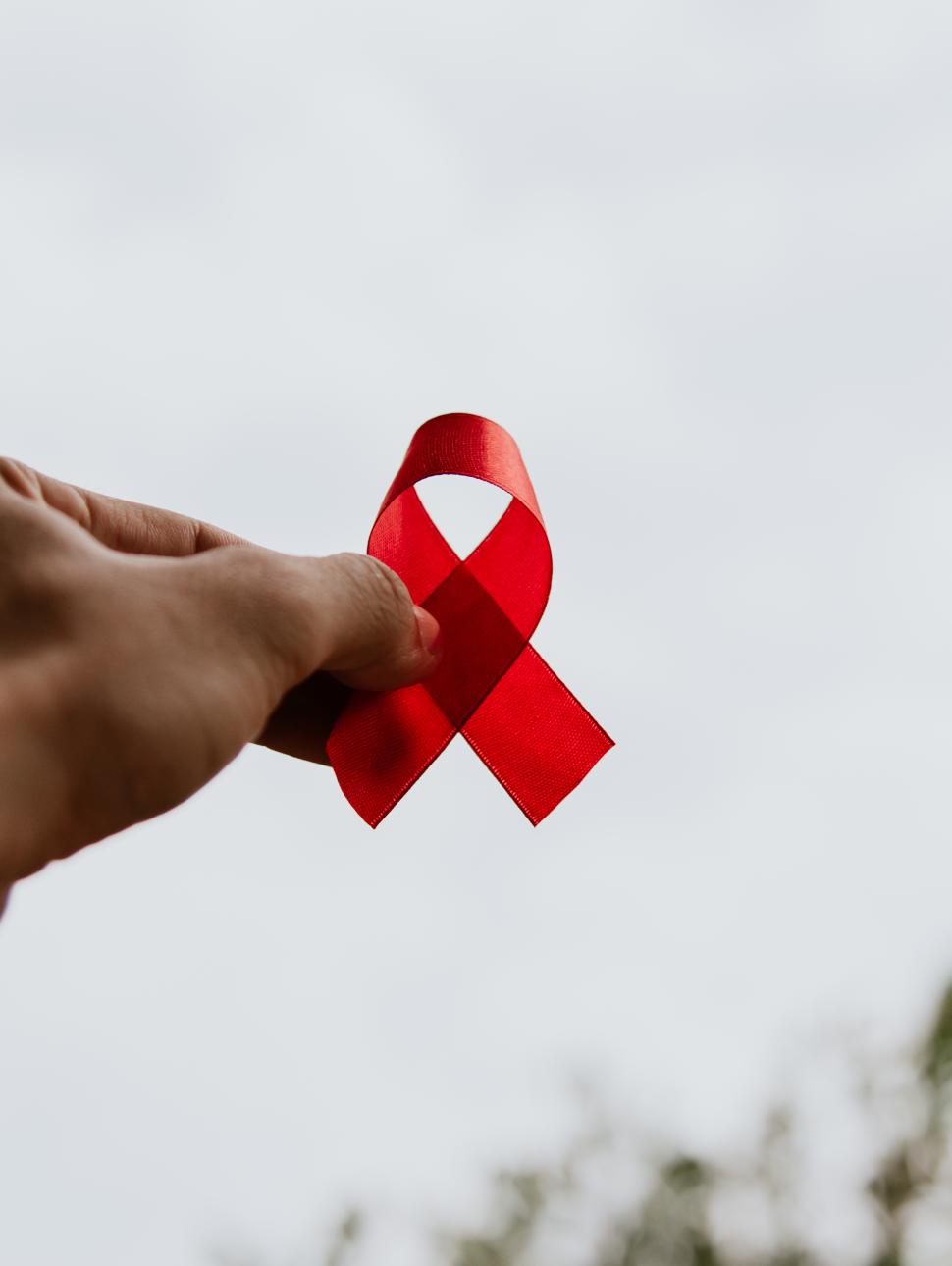 A hand holds a red ribbon folded over itself up against a bright sky. The red ribbon is symbolic for the World Aids Day awareness campaign.