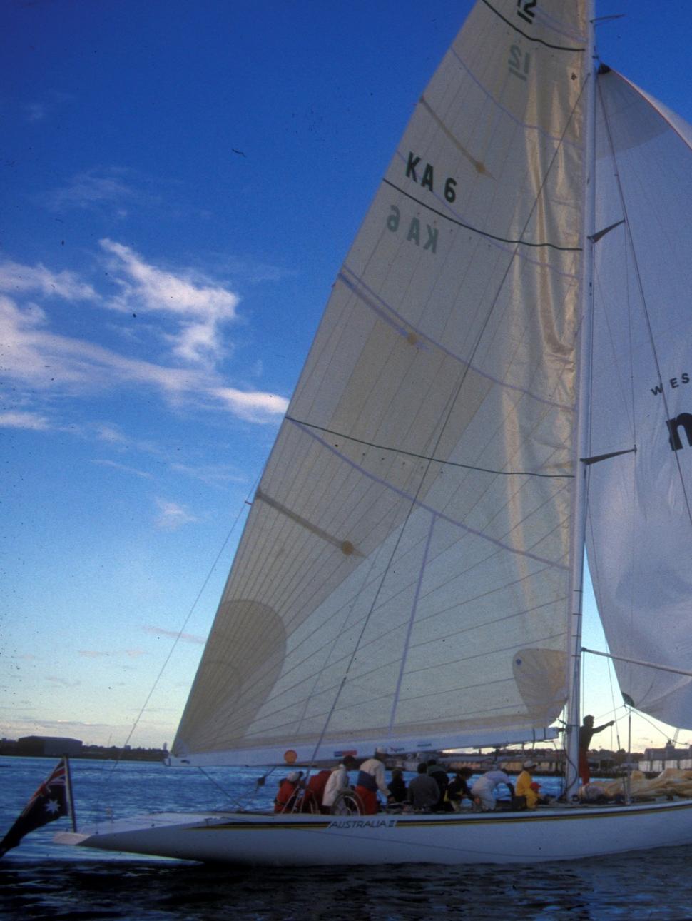 The Australia II yacht, with a long off-white sail, and a WA Museum branded sail, moves towards the Maritime Museum in Fremantle which appears to be underconstruction, dating the image to the late 1990s or early 2000s