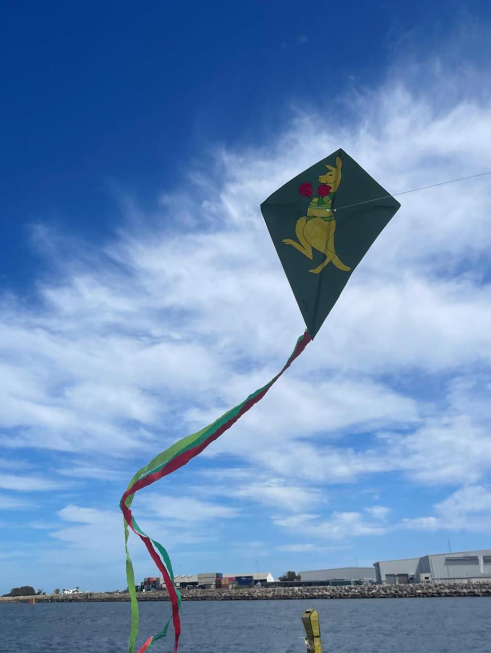 Green kite flying with a yellow boxing kangaroo printed. the kite has streamer tails and the sky is blue with clouds in the background