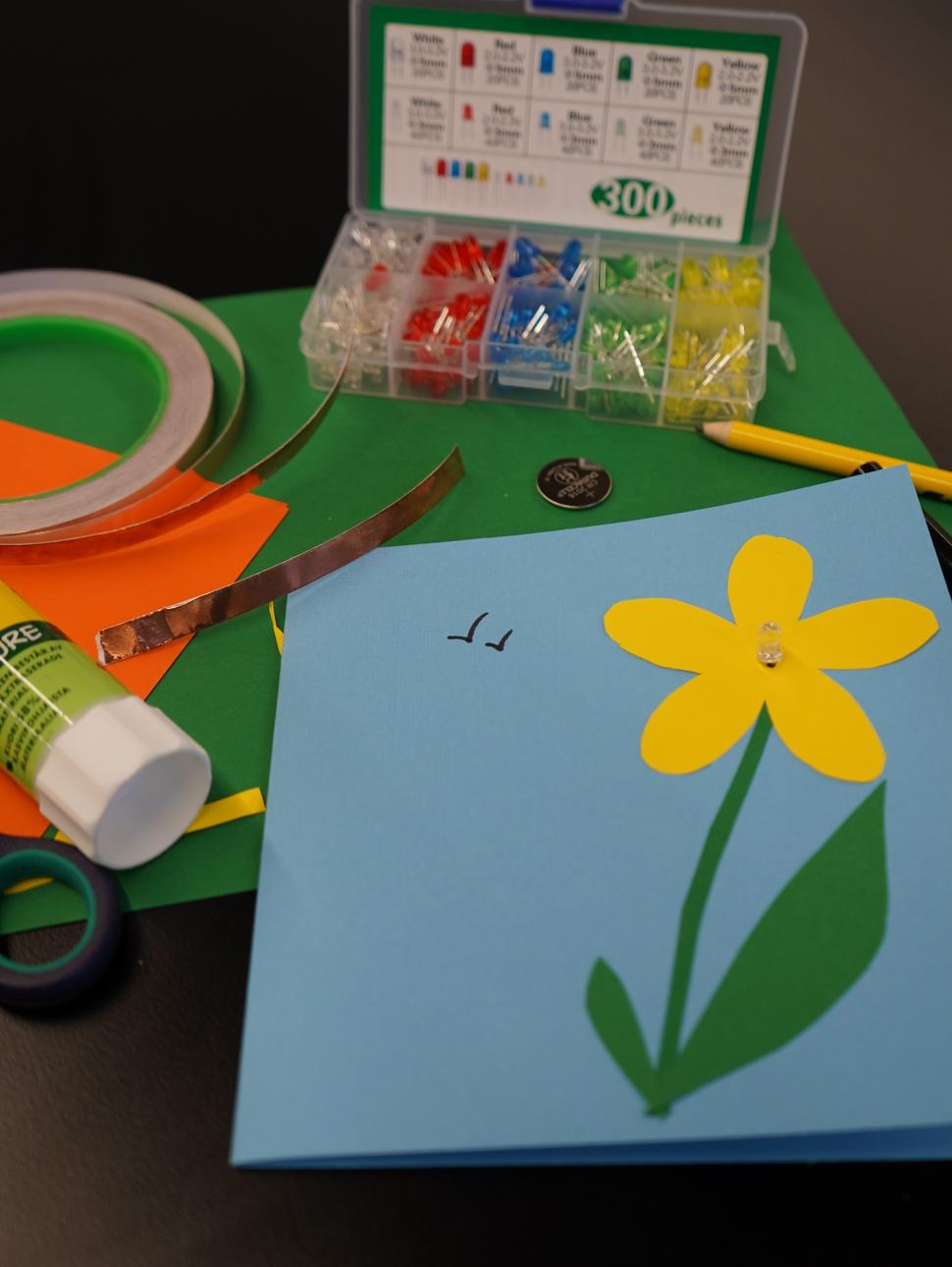 The image shows a hand made cars with a yellow flower on t blue background. Behind i are all of th items required to turn this into a card that lights up using an electrical circuit.