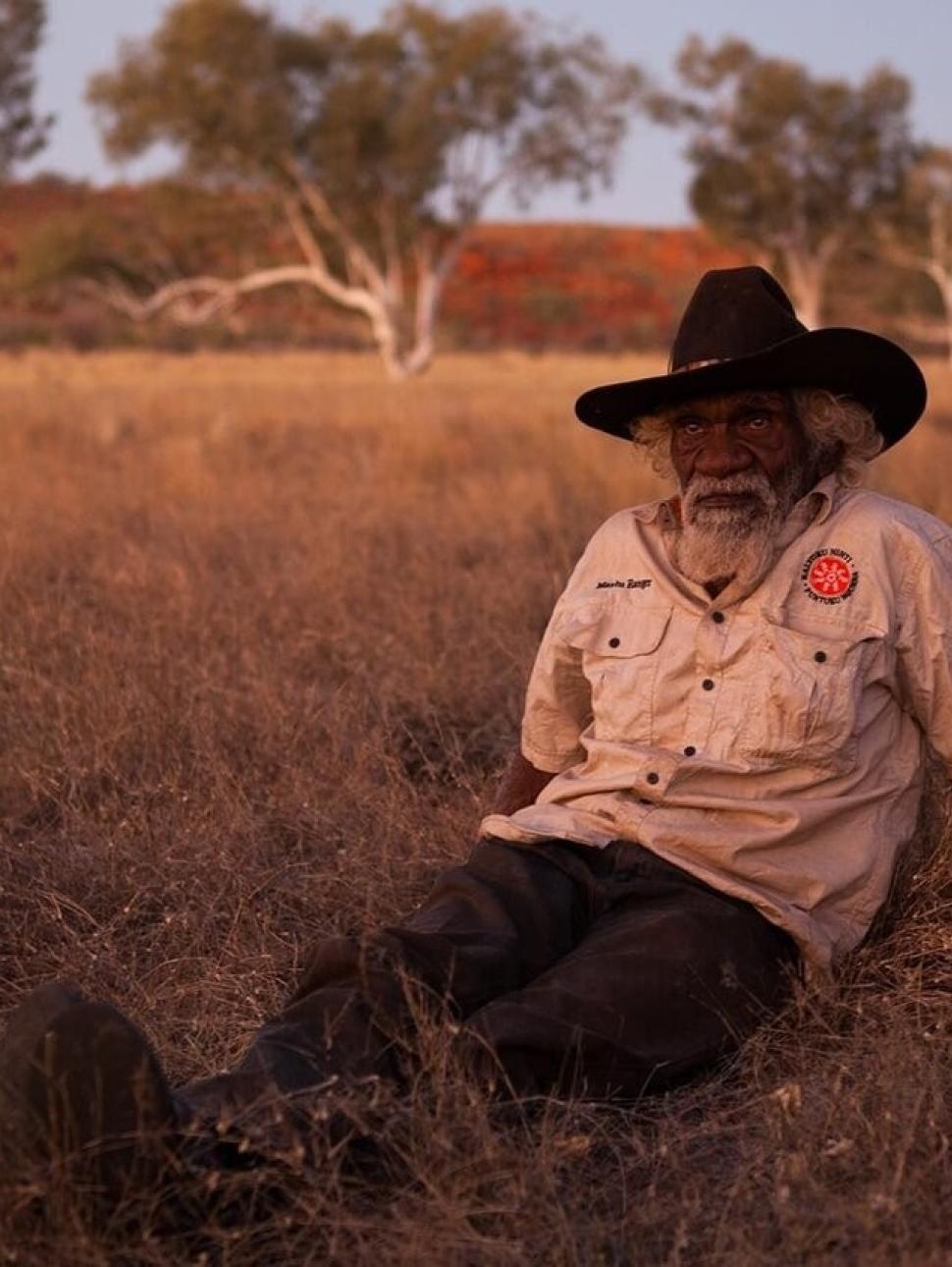 Aboriginal elder Nyarri Nyarri Morgan wears a khaki coloured ranger shirt and a black stockmans hat and sits out on country at dusk, with the trees and grass beginning to darken behind him.