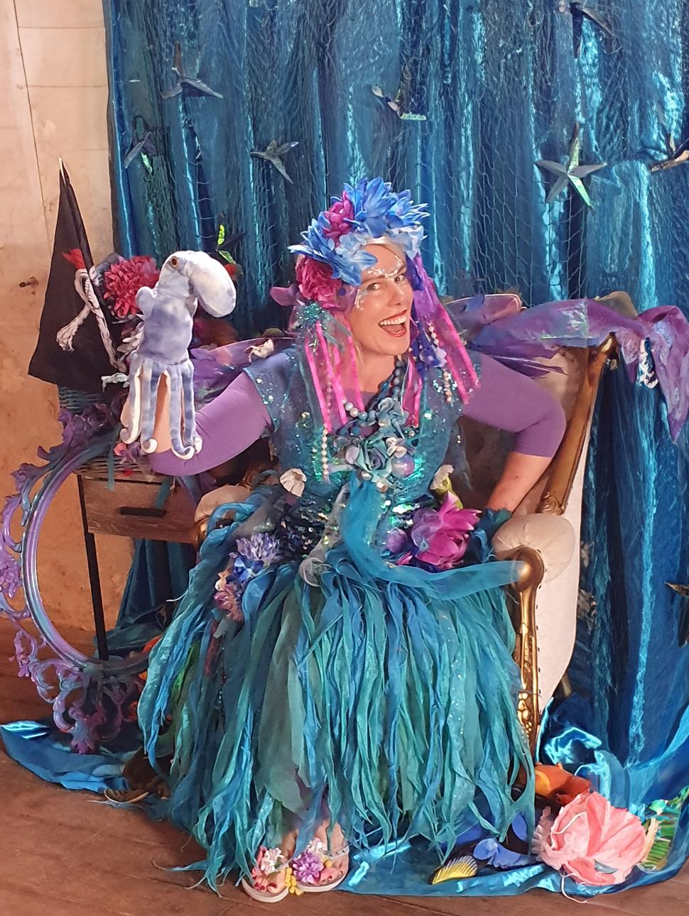 a person with blue hair and blue green clothes looking like an underwater scene sits on a purple chair