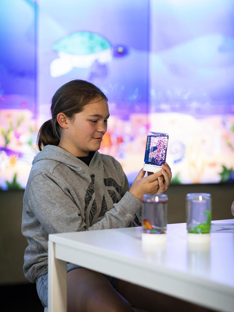a child sits at a table and looks at a home made snow globe. it has small sparkles swirling inside the globe. other materials are on the table