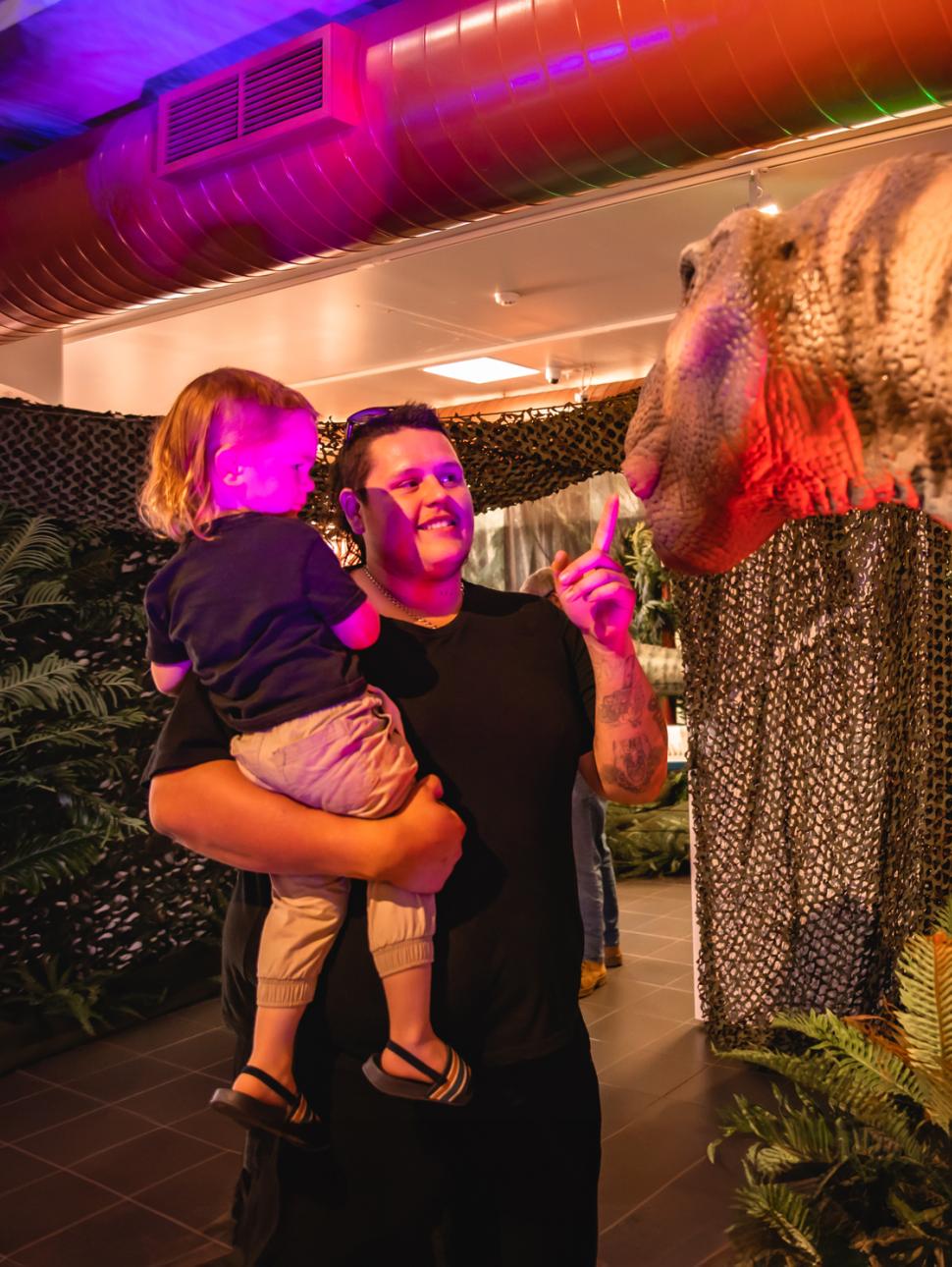 an adult carries a child and they both look to a dinosaur animatronic on display with vegetation and lights in the immediate surrounds