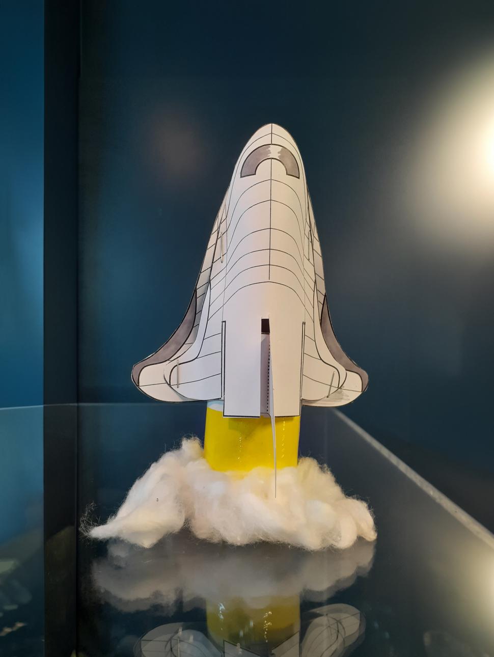 A paper space craft with cotton wool at the base sits on a table with blue walls in the background